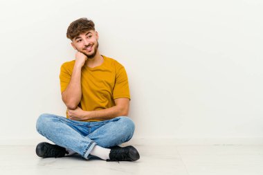 Faslı genç bir adam beyaz arka planda tek başına oturmuş gülümsüyor ve kendinden emin, elleriyle çenesine dokunuyor..