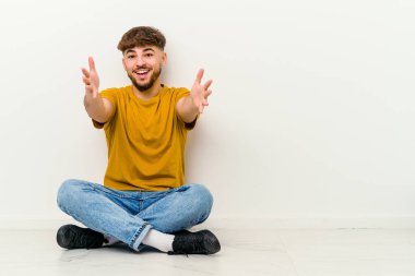 Beyaz arka planda izole bir şekilde yerde oturan Faslı genç adam kameraya sarıldığı için kendini güvende hissediyor..