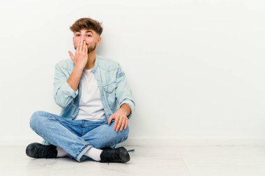 Faslı genç bir adam beyaz arka planda izole bir şekilde oturuyor. Şok olmuş, ağzı ellerle kaplanmış, yeni bir şey keşfetmek için sabırsızlanıyor..