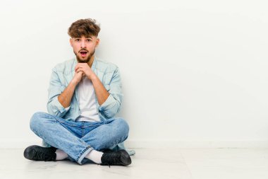 Faslı genç bir adam beyaz arka planda izole bir şekilde yerde oturmuş şans diliyor, şaşırmış ve ağzını açıp önüne bakıyor..