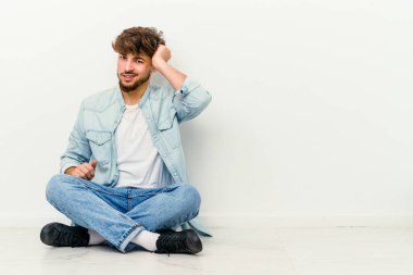 Genç Faslı bir adam beyaz arka planda izole bir şekilde yerde oturmuş zaferi, tutkuyu ve coşkuyu kutluyor, mutlu bir ifade..