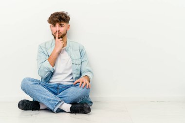 Genç Faslı bir adam beyaz arka planda izole bir şekilde oturuyor. Düşünüyor, düşünüyor, düşünüyor, hayal kuruyor..