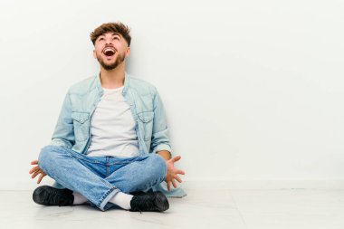 Genç Faslı bir adam beyaz arka planda izole bir şekilde yerde oturmuş kızgın, öfkeli ve hayal kırıklığına uğramış bir şekilde bağırıyor..