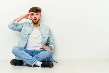 Faslı genç bir adam beyaz arka planda izole bir şekilde yerde oturuyor. Uzağa bakıyor. Alnını tutuyor..