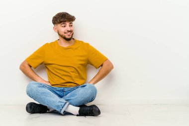 Faslı genç bir adam beyaz arka planda izole bir şekilde yerde oturuyor mutlu bir şekilde gülüyor ve ellerini karnının üstünde tutarak eğleniyor..