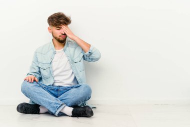 Faslı genç bir adam beyaz arka planda izole bir şekilde yerde oturuyor. Başı ağrıyor, yüzüne dokunuyor..