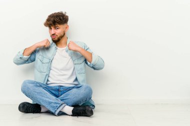 Genç Faslı bir adam beyaz arka planda izole bir şekilde yerde oturuyor bir tartışma yüzünden yumruk, öfke, kavga atıyor, boks yapıyor..