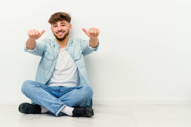 Faslı genç bir adam beyaz arka planda izole bir şekilde yerde oturuyor. Baş parmağını kaldırıyor, gülümsüyor ve kendinden emin..