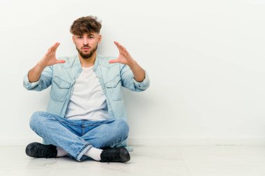 Faslı genç bir adam beyaz arka planda izole bir şekilde yerde oturuyor. Elinde avuç içi bir şeyle kameraya sunuyor..