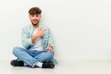 Faslı genç bir adam beyaz arka planda izole bir şekilde oturuyor gülümsüyor ve boş alanda bir şeyler gösteriyor..