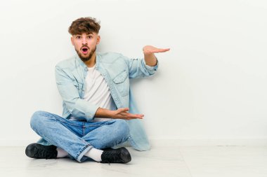 Beyaz arka planda izole bir şekilde yerde oturan genç Faslı adam elleri arasında bir kopya alanı tutarken şok ve hayrete düştü..