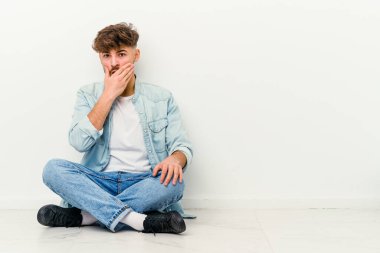 Genç Faslı bir adam beyaz arka planda izole bir şekilde yerde oturuyor. Ağzı kapalı ve elleri endişeli görünüyor..