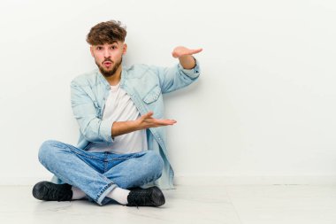 Yerde oturan genç Faslı adam beyaz arka planda izole bir şekilde iki eliyle bir şey tutuyor, ürün sunumu..