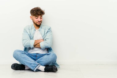 Genç Faslı adam beyaz arka planda izole bir şekilde yerde oturuyor. Kollarını kavuşturup gülümsüyor..