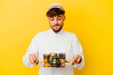 Genç Faslı adam tipik Arap kostümünü giyiyor sarı arka planda izole edilmiş çay içiyor.