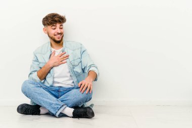 Genç Faslı bir adam beyaz arka planda izole bir şekilde yerde oturuyor. Gülüyor, elini kalbe koyuyor, mutluluk kavramını koruyor..