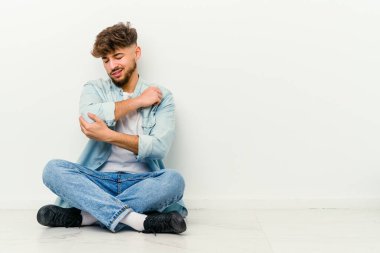 Faslı genç bir adam beyaz arka planda yalnız başına oturuyor dirsek masajı yapıyor, kötü bir hareketin ardından acı çekiyor..