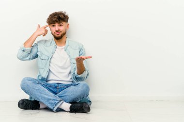 Faslı genç bir adam beyaz arka planda izole bir şekilde yerde oturuyor ve elinde bir ürün tutuyor..