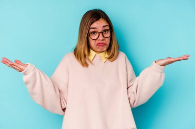 Genç beyaz kadın, mavi arka planda izole edilmiş. Sorgulama sırasında omuz silkiyor ve şüphe ediyor..
