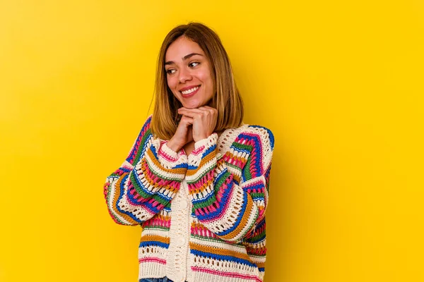 Young caucasian skinny woman isolated on yellow background keeps hands under chin, is looking happily aside.