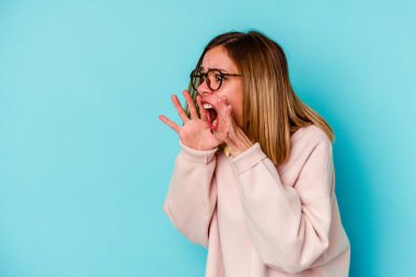 Mavi arka planda izole edilmiş genç beyaz kadın yüksek sesle bağırıyor, gözleri açık ve elleri gergin tutuyor..
