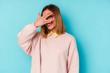 Mavi arka planda izole edilmiş genç beyaz kadın kameraya parmaklarıyla göz kırpıyor, yüzünü kapatmaktan utanıyor..