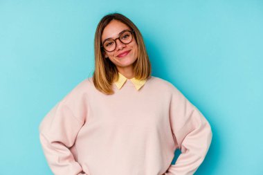 Mavi arka planda izole edilmiş genç beyaz kadın ellerini kalçasında tutuyor..