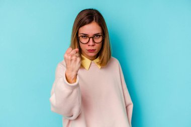 Mavi arka planda izole edilmiş genç beyaz kadın yumruğunu kameraya gösteriyor, agresif yüz ifadesi..