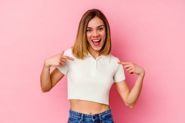 Pembe arka planda soyutlanmış beyaz bir kadın. Parmakla göstererek şaşırmış, geniş bir gülümsemeyle..