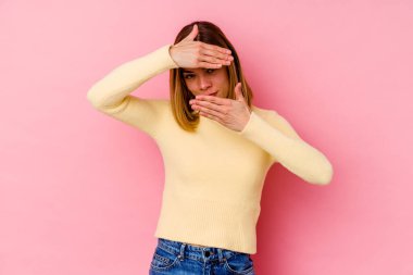 Pembe arka planda izole edilmiş genç beyaz kadın kameraya parmaklarıyla göz kırpıyor, yüzünü kapatmaktan utanıyor..