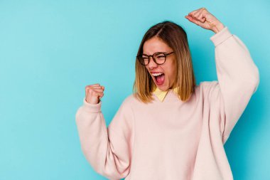 Mavi arka planda izole edilmiş genç beyaz kadın zafer kazandıktan sonra yumruğunu kaldırıyor, kazanan konsept.