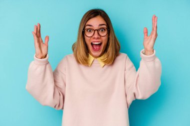 Mavi arka planda izole genç beyaz kadın hoş bir sürpriz alıyor, heyecanlı ve el kaldırıyor..