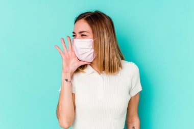 Mavi arka planda izole edilmiş virüs maskesi takan genç bir kadın bağırıyor ve avuç içini açık tutuyor..