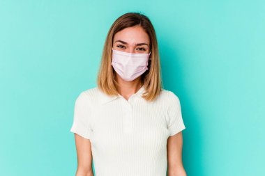 Mavi arka planda izole edilmiş virüs maskesi takan genç bir kadın mutlu, güler yüzlü ve neşeli..