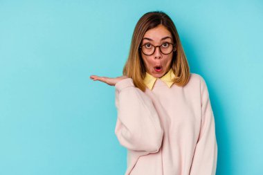 Mavi arka planda izole edilmiş genç beyaz kadın avuç içi fotokopi alanını elinde tutmaktan etkilenmiş..