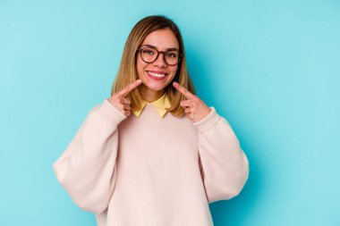 Mavi arka planda izole edilmiş genç beyaz kadın gülümsüyor, parmaklarını ağıza doğrultuyor..