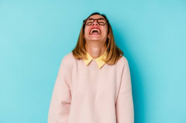 Genç beyaz bir kadın öğrenci mavi arka planda izole edilmiş rahat ve mutlu bir şekilde gülüyor, boynunu uzatmış dişlerini gösteriyor..