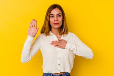 Beyaz tenli genç kadın sarı arka planda izole edilmiş yemin ediyor, elini göğsüne koyuyor..