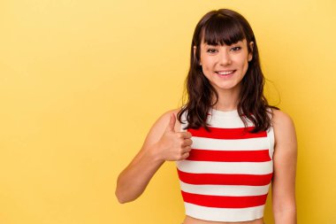 Sarı arka planda izole edilmiş genç beyaz kadın gülümsüyor ve başparmağını kaldırıyor.