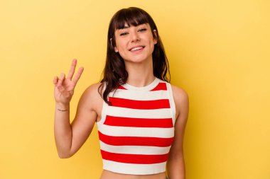 Sarı arka planda izole edilmiş genç beyaz kadın zafer işareti gösteriyor ve gülümsüyor..