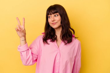 Sarı arka planda izole edilmiş genç beyaz kadın neşeli ve kaygısız parmaklarıyla barış sembolü gösteriyor..