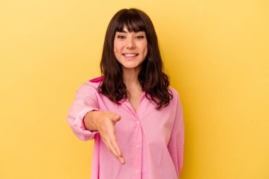 Sarı arka planda izole edilmiş genç beyaz kadın selamlaşma jesti olarak kameraya elini uzatıyor..