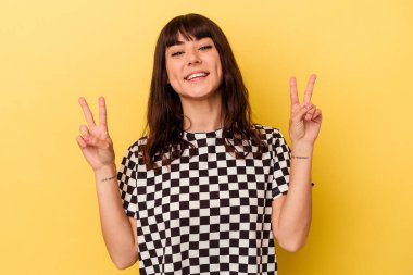 Sarı arka planda izole edilmiş genç beyaz kadın zafer işareti gösteriyor ve gülümsüyor..