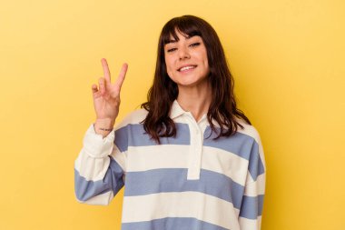 Sarı arka planda izole edilmiş genç beyaz kadın neşeli ve kaygısız parmaklarıyla barış sembolü gösteriyor..