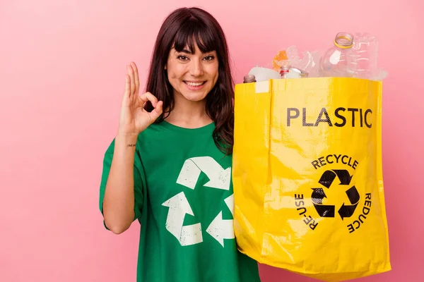 年轻的高加索女人拿着一个用粉红背景隔开的回收塑料袋 快乐而自信地做出了一个好的姿态 — 图库照片