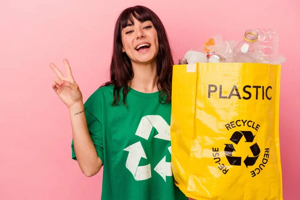 年轻的高加索女人拿着一个回收的塑料袋 与粉色背景隔离 快乐而无忧无虑 用手指展示和平的象征 — 图库照片