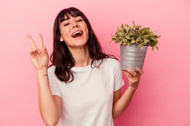 Genç beyaz kadın pembe arka planda izole edilmiş bir bitki tutuyor neşeli ve kaygısız parmaklarıyla bir barış sembolü gösteriyor..