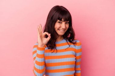 Pembe arka planda izole edilmiş genç beyaz kadın bir göz kırpıyor ve elinde iyi bir el hareketi tutuyor..