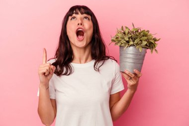 Genç beyaz kadın pembe arka planda izole edilmiş bir bitki tutuyor. Ağzı açık bir şekilde üst tarafı işaret ediyor..