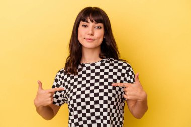 Sarı arka planda izole edilmiş genç beyaz bir kadın gömleğin fotokopi odasını elle işaret ediyor, gururlu ve kendinden emin.